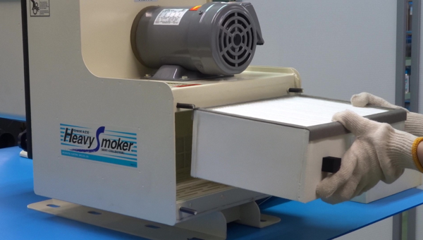 ヘビースモーカー（HVS）ミストコレクターのフィルター交換作業。前面から簡単に取り外しでき、メンテナンス性の高さを示している。